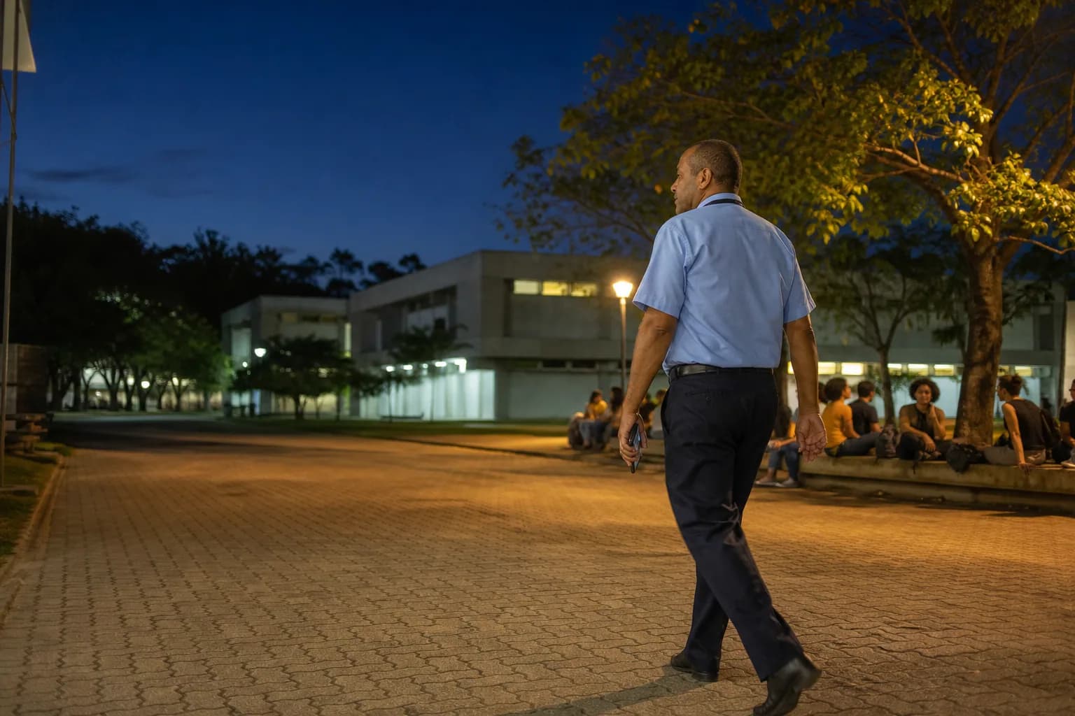Início da noite no calçadão de um campus universitário brasileiro: um trabalhador de uniforme claro percorre a rota com o celular ao lado do corpo, enquanto estudantes conversam ao fundo sob uma árvore.